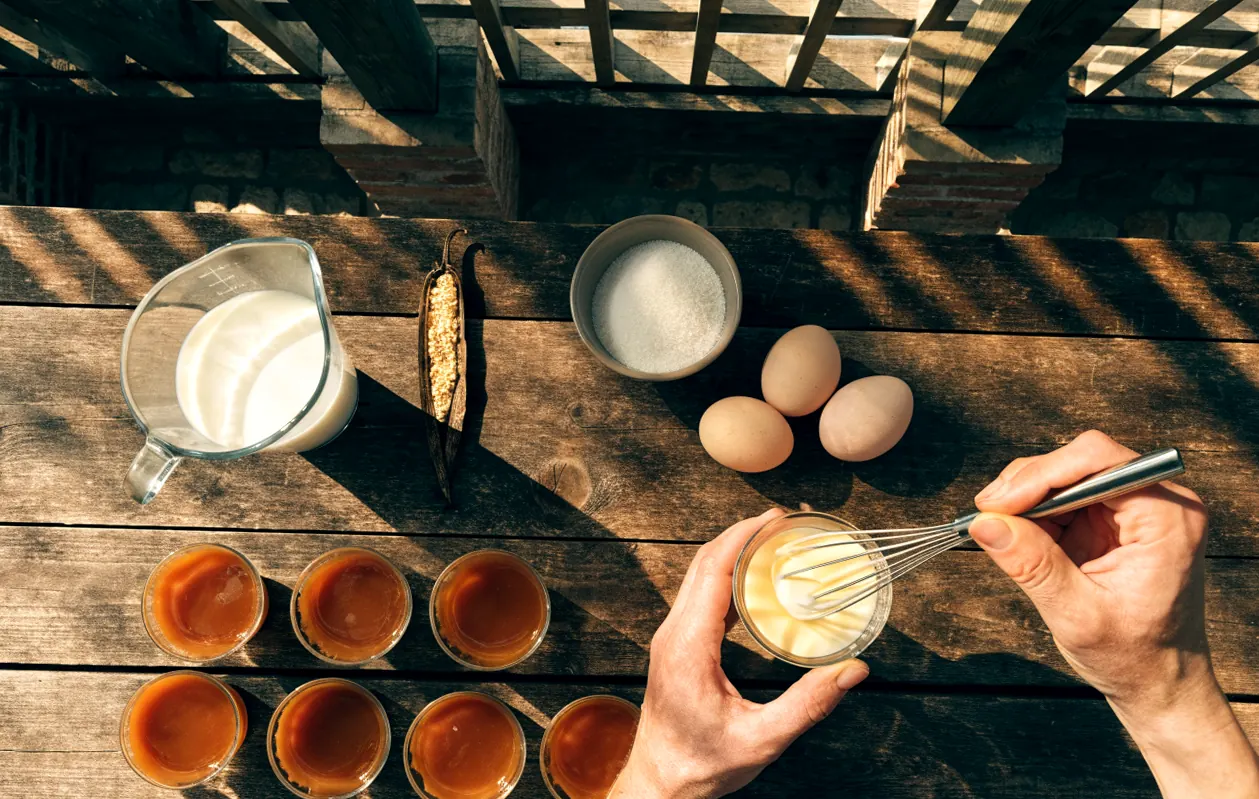 Verre doseur rempli d’un demi-litre de lait, œufs, vanille et ramequins caramélisés sur une table rustique